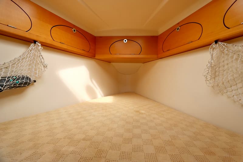 De kajuit toont een V-vormig bed met een lichtgekleurd matras, omringd door houten kasten en twee netten aan de wanden.
