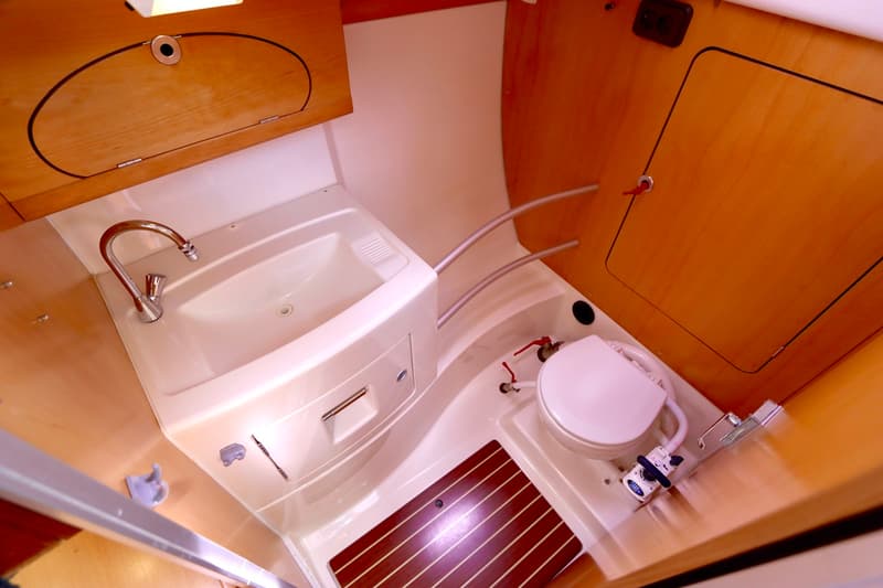 Een boottoiletruimte met een wasbak, kraan, toilet, houten kasten en een houten vloerrooster is zichtbaar.