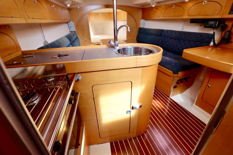 Een bootinterieur met een kombuisblok met spoelbak en kooktoestel, omringd door houten kasten en blauwe zitbanken, en een…