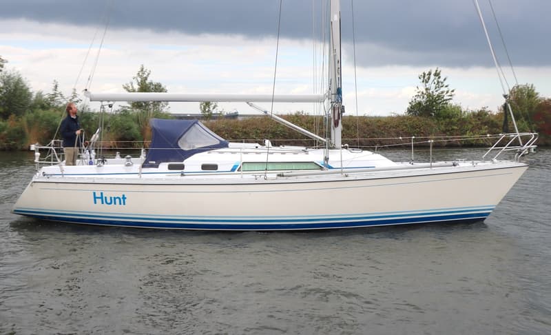 Een witte zeilboot met blauwe strepen op de romp en de naam "Hunt" vaart op het water, met een persoon aan het roer.