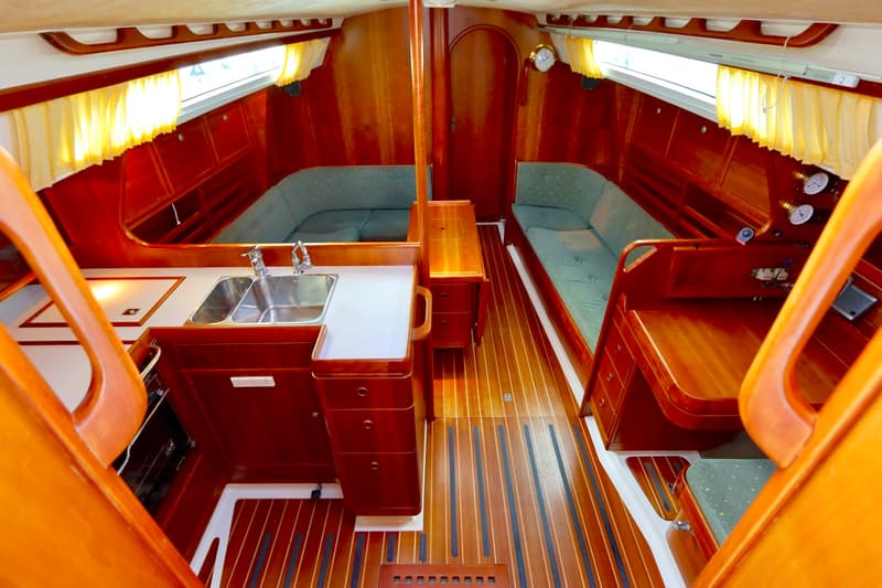 De foto toont een houten bootinterieur met een kombuis, twee banken met groene kussens en een houten vloer.