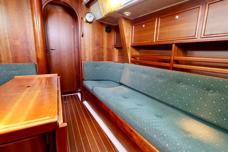 De foto toont een houten bootinterieur met een tafel, groene bankkussens, een deur, kasten en een klok aan de wand.