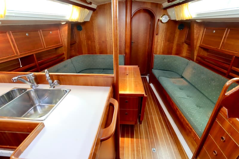 De foto toont een houten kajuitinterieur met een kombuis met dubbele spoelbak en twee groene gestoffeerde banken.
