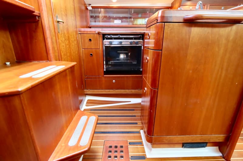Een houten bootinterieur toont een kombuis met een ingebouwde oven, kasten, laden en een houten vloer met treden.