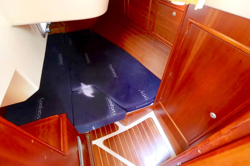 De foto toont een kajuitinterieur met donkerblauwe matrassen, houten wanden en een houten vloer met een wit rooster.