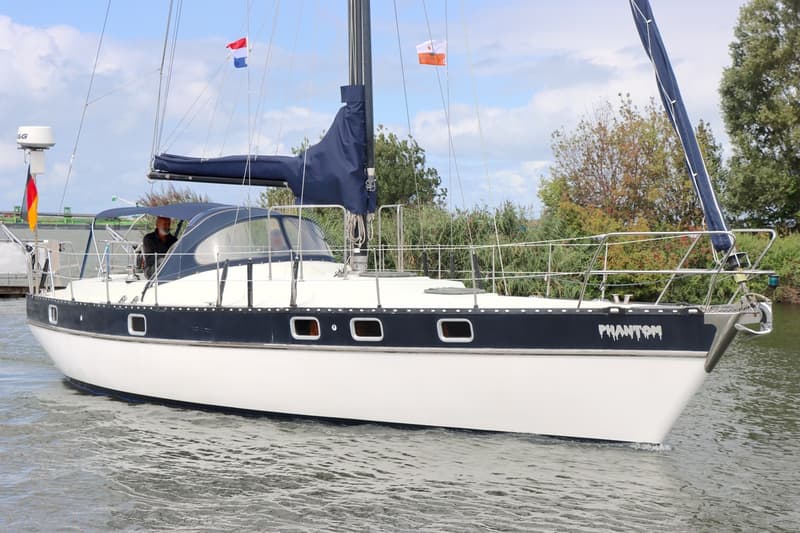 Een witte zeilboot met donkerblauwe band en de naam "PHANTOM" vaart op het water, met een persoon in de kuip en vlaggen.