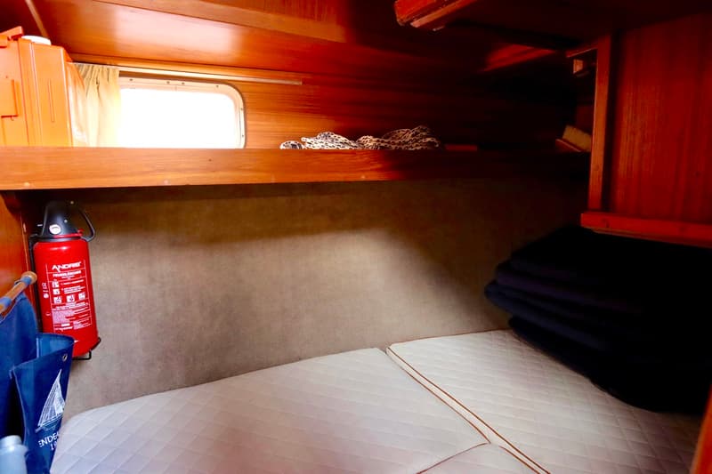 De kajuit toont houten betimmering, een raam met gordijn, een rode brandblusser, witte matrassen en een oranje kist.