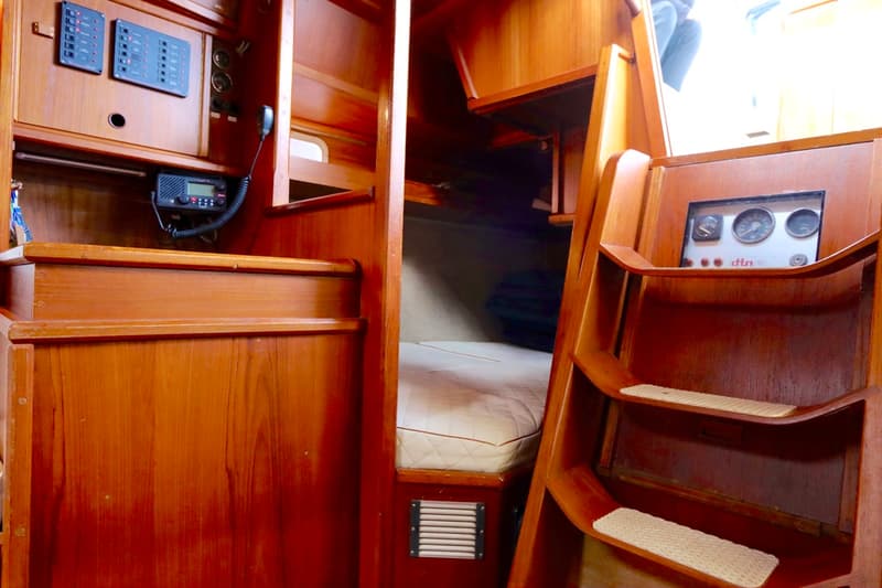 De foto toont een houten bootinterieur met een bed, een marifoon, schakelpanelen, meters en een trap.