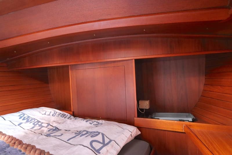 Een houten kajuitinterieur met een bed, opbergvakken, een plafond met inbouwspots en een klein elektrisch kastje.