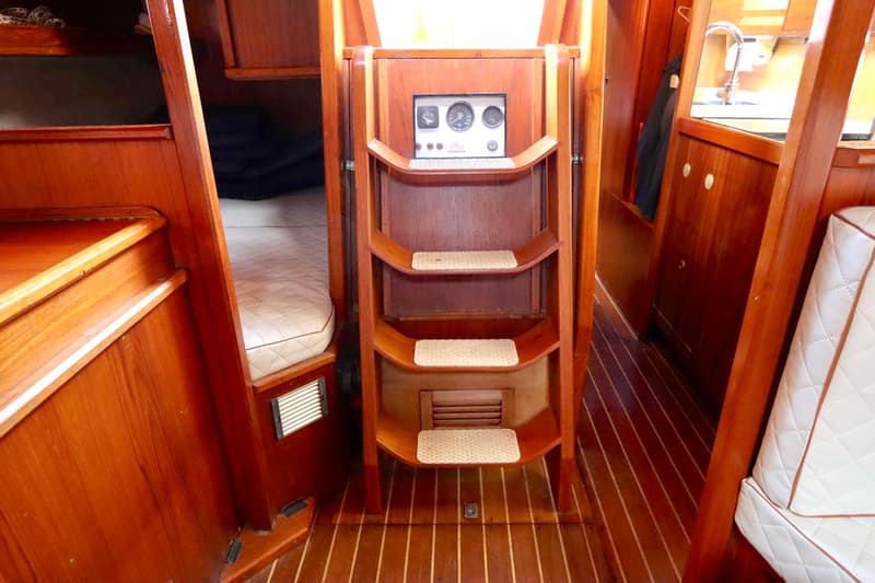 Een houten scheepstrap met drie treden en een instrumentenpaneel erboven is zichtbaar in een houten interieur.