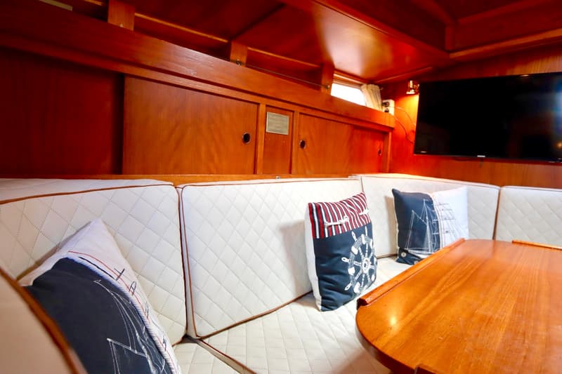 Een houten interieur met witte gestikte kussens, decoratieve kussens, een houten tafel en een televisie is zichtbaar.