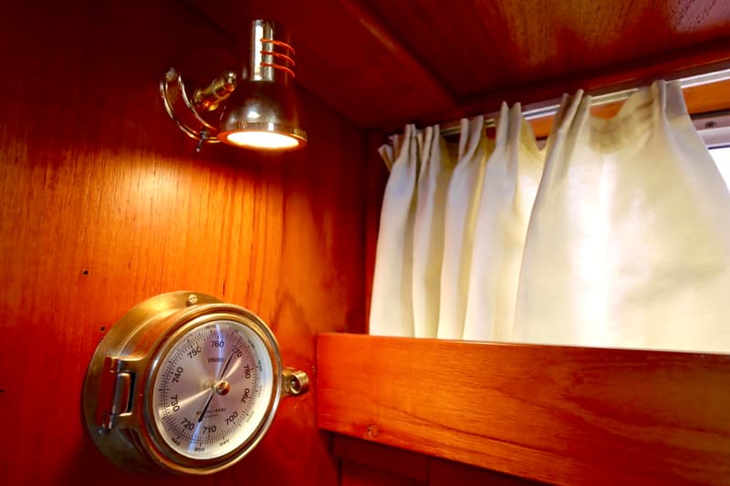 Een messingkleurige lamp en een barometer zijn gemonteerd op een houten wand, met lichte gordijnen ernaast.