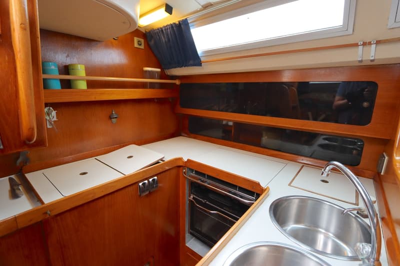 De kombuis toont een houten interieur met een wit aanrechtblad, dubbele spoelbak, kraan, ingebouwde oven en opbergplanken.