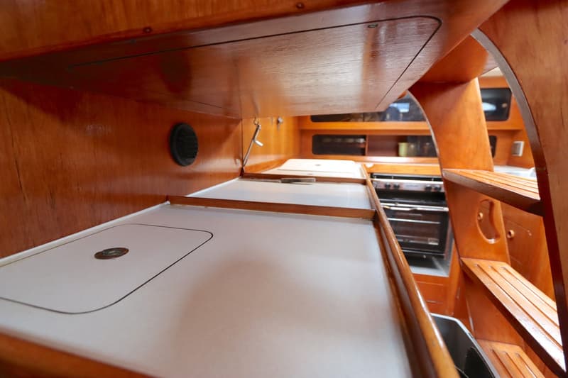 De foto toont een houten kajuitinterieur met een wit aanrechtblad, een zwart ventilatierooster en een fornuis/oven in de…