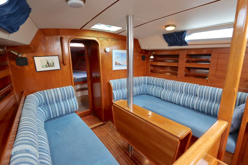 De kajuit toont een houten interieur met blauwe zitkussens en gestreepte rugkussens, een opklapbare tafel en een doorgang…