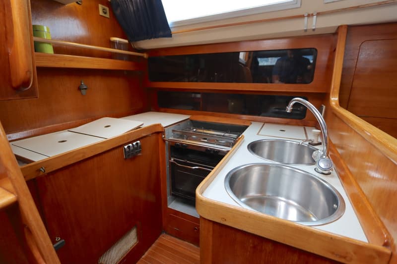 Een houten kajuitinterieur met een kombuis voorzien van twee RVS spoelbakken, een kraan en een gasfornuis met oven.