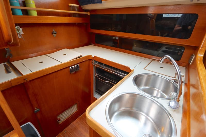 De kombuis toont een houten interieur met witte werkbladen, twee roestvrijstalen spoelbakken, een kraan en een oven.