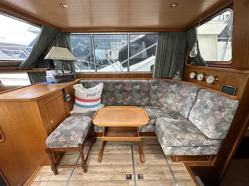 Een interieur van een boot met een U-vormige bank met patroon, een houten tafel en een kussen met de tekst "Pavasas post".
