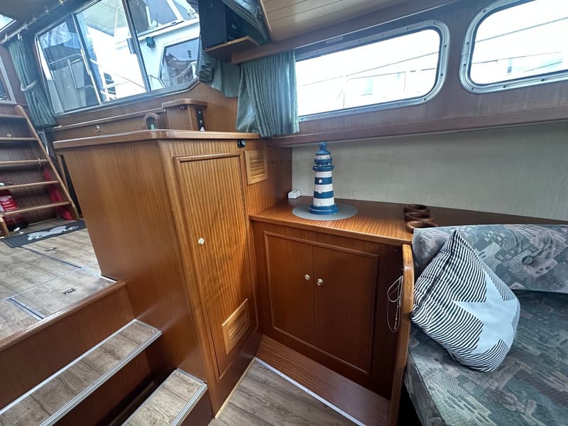Een interieur van een boot met houten lambrisering, ramen, een bank met kussens en een decoratieve vuurtoren.