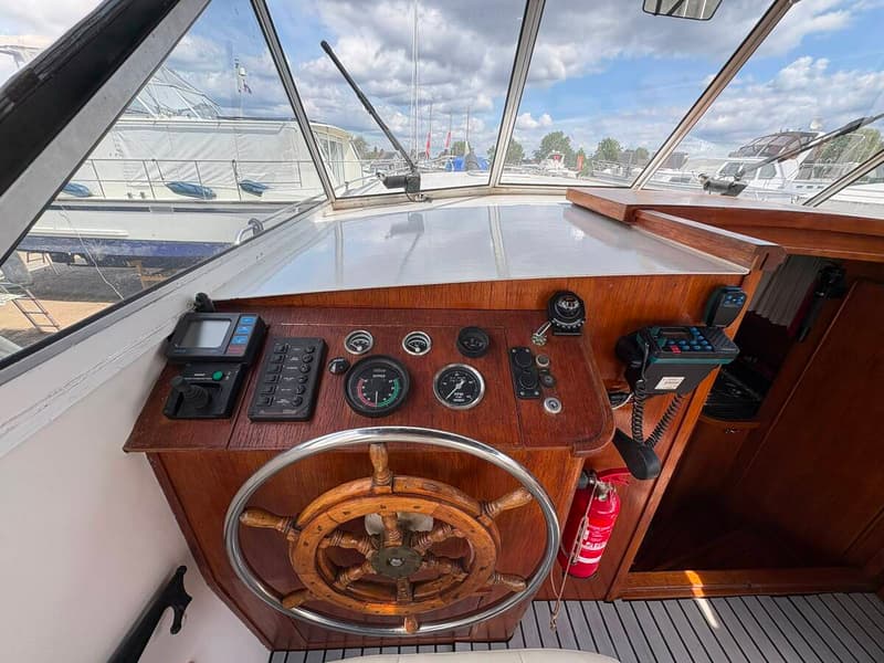 Een houten stuurwiel en dashboard met diverse meters, knoppen en navigatieapparatuur zijn zichtbaar.