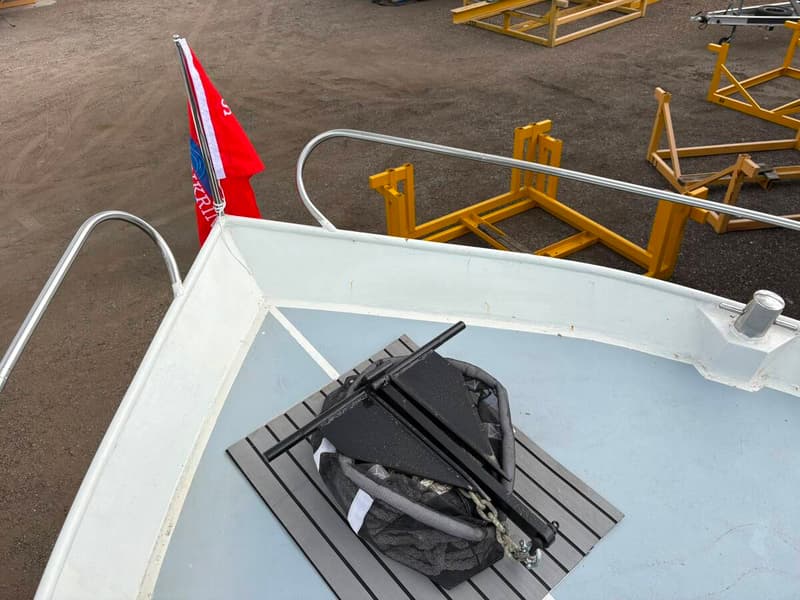 Een zwart anker ligt op een grijze mat op het dek van een boot, met een rode vlag aan de boeg. — Valk Sport