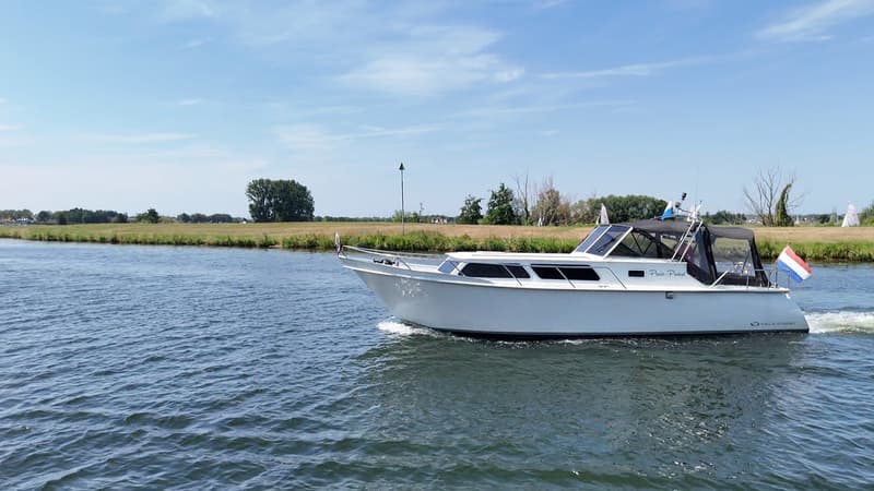 Een witte motorboot met een Nederlandse vlag vaart op een waterweg met groene oevers onder een blauwe lucht. — Valk Sport