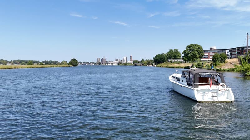 Een witte motorboot met een zwarte kap vaart op een rivier met een stad op de achtergrond. — Valk Sport