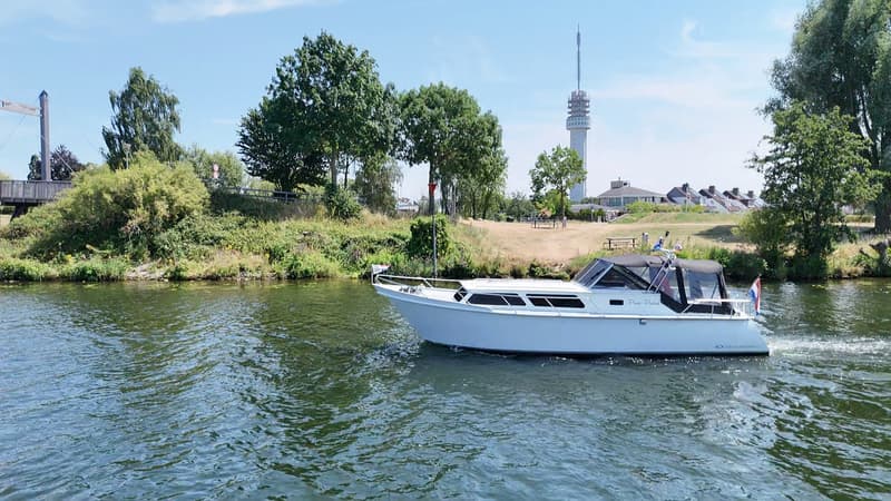 Een witte motorboot met een zwarte kap vaart over het water, met een brug en een zendmast op de achtergrond. — Valk Sport
