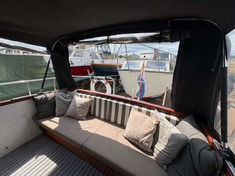 Een bootinterieur met een bank met kussens en een gestreepte rugleuning, met uitzicht op andere boten.
