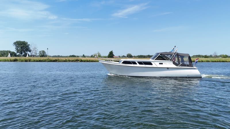Een witte motorboot met een zwarte kap vaart op een waterweg met een groene oever en blauwe lucht. — Valk Sport