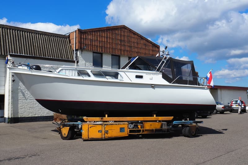 Een witte boot met de naam "PASSE-PARTOUT" staat op een gele trailer voor een gebouw met een pannendak. — Valk Sport