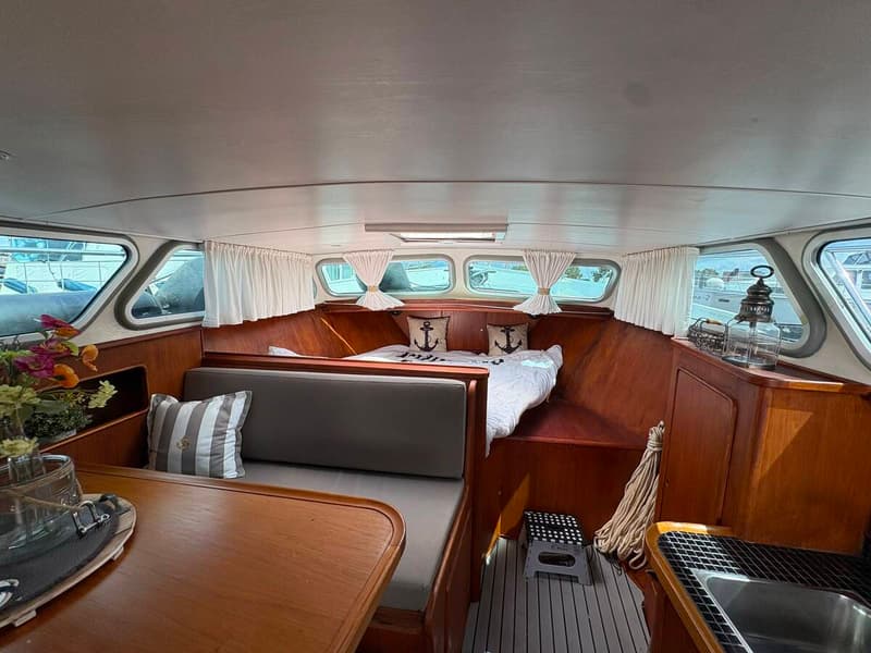De foto toont het interieur van een boot met een houten tafel, banken, een bed, ramen met gordijnen en een gootsteen.