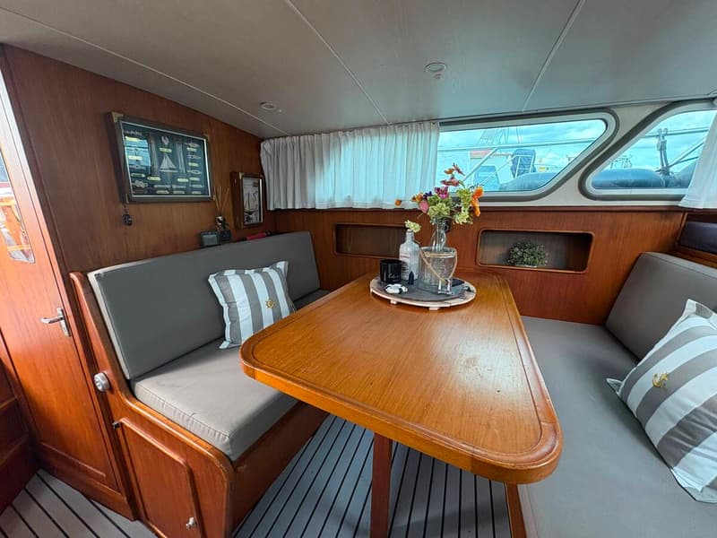 De foto toont een kajuit met een houten tafel, grijze banken met kussens, en ramen met witte gordijnen. — Valk Sport