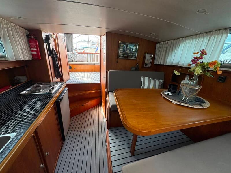 De foto toont het interieur van een boot met een kombuis, eettafel en zitbanken, en een trap naar een open achterdek.