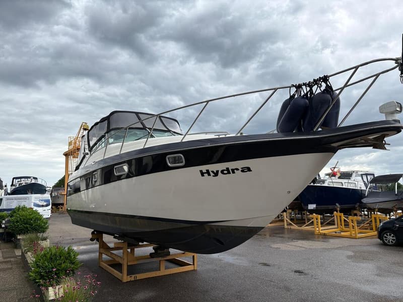 Een witte boot met zwarte accenten en de naam "Hydra" op de boeg staat op een houten bok onder een bewolkte hemel.