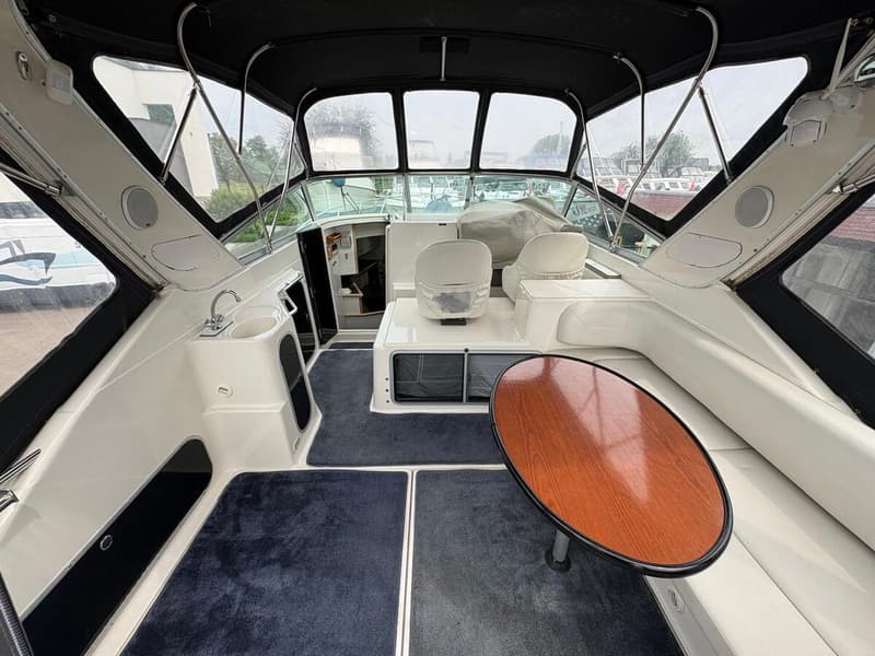 De foto toont het interieur van een boot met een donkerblauw tapijt, witte banken en een bruine ovale tafel. — Maxum 3200 SCR