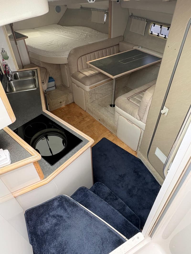 De foto toont het interieur van een boot met een bed, een zithoek met tafel, een keukenblok met spoelbak en kookplaat, en…