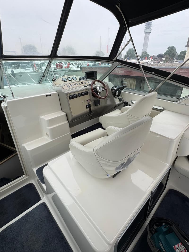 De foto toont de cockpit van een boot met een stuurwiel, instrumentenpaneel, twee witte stoelen en een uitzicht op andere…