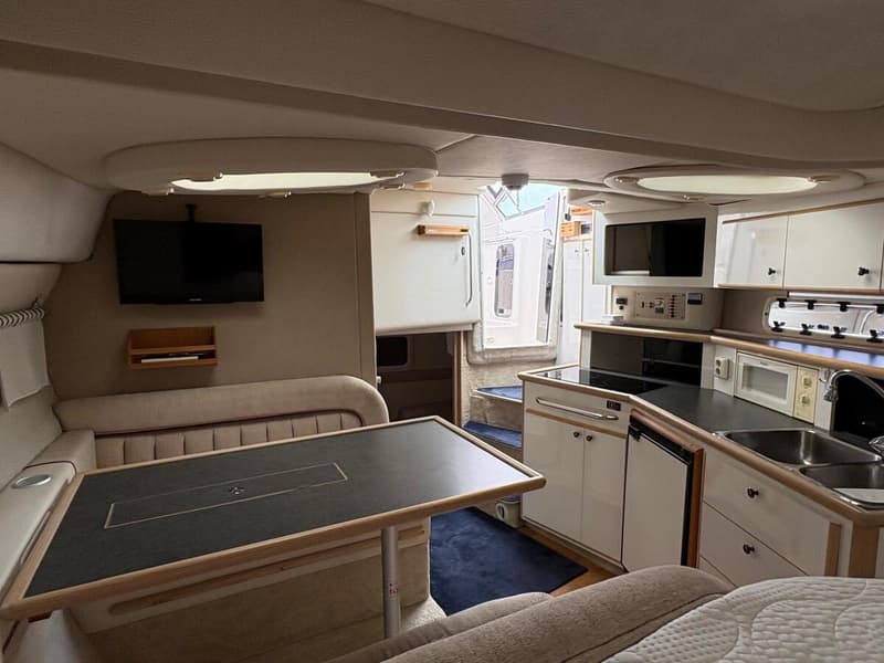 De foto toont het interieur van een boot met een zithoek, tafel, televisie, keukenblok en een doorgang naar buiten.