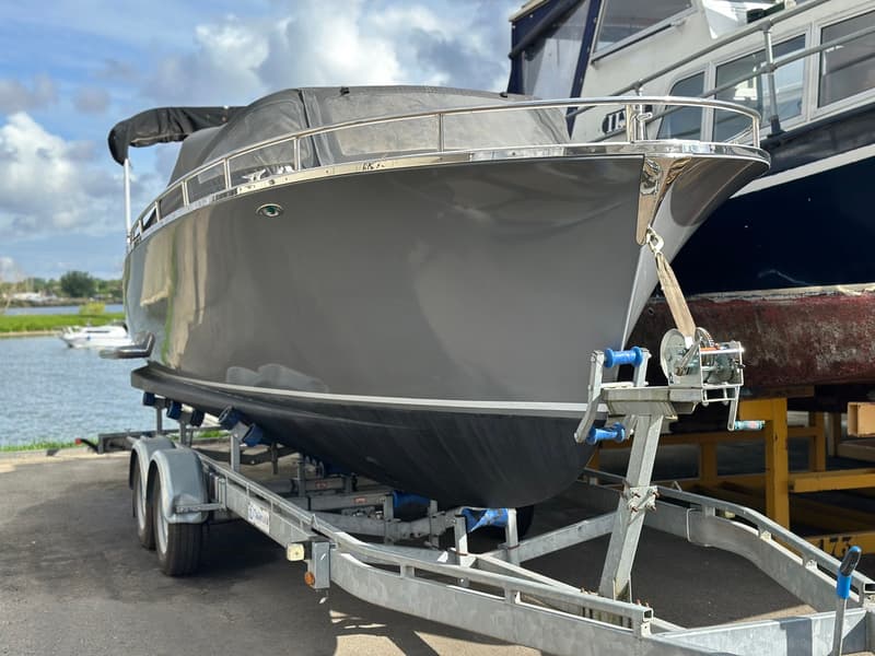 Een grijze boot met zwarte onderkant staat op een trailer, met een lier aan de voorkant, bij een waterkant. — Riviera 24