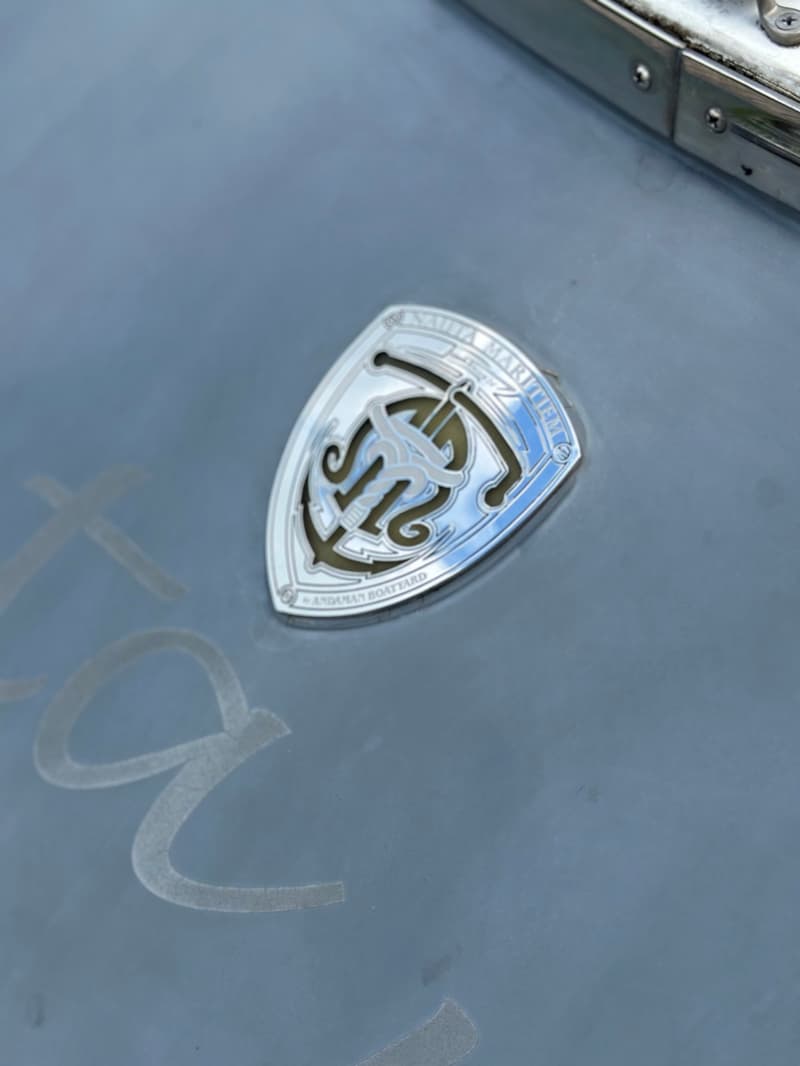 Een close-up van een zilverkleurig embleem met een anker en de tekst "ANDAMAN BOATYARD" op een grijze ondergrond.