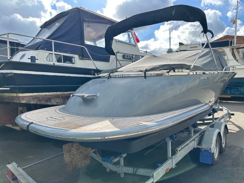 Een grijze boot met een zwarte kap en een afdekzeil staat op een trailer, met een donkerblauwe boot erachter. — Riviera 24