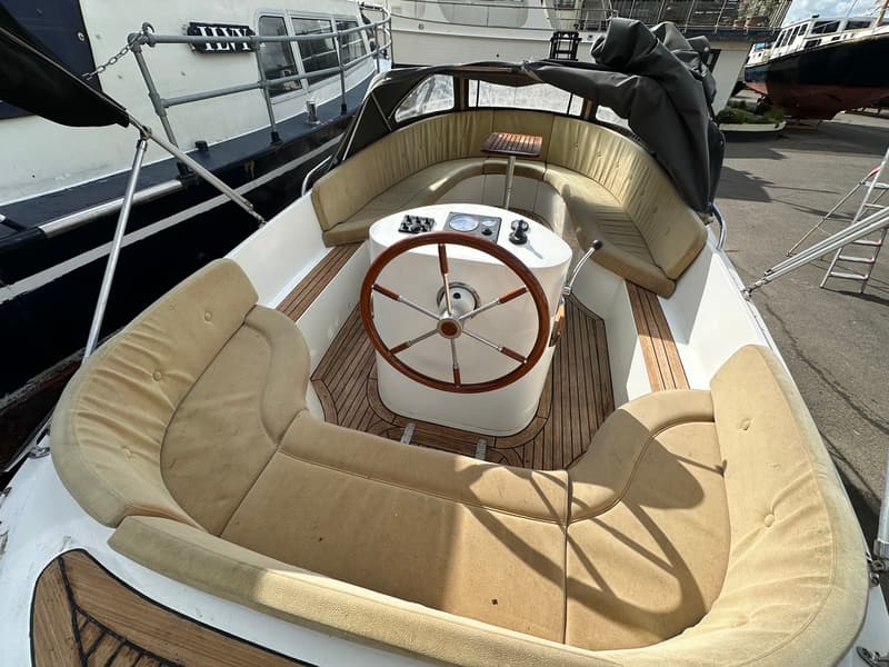 De foto toont de cockpit van een boot met een houten stuurwiel, beige kussens en een houten vloer. — Riviera 24
