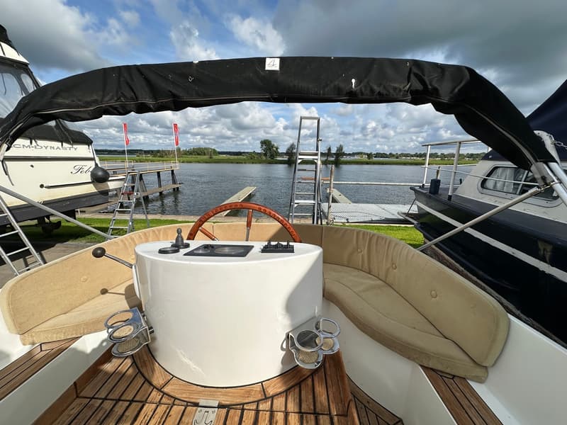 Een bootinterieur met een stuurwiel, beige kussens, een houten vloer en een zwart afdak, met uitzicht op water en andere…