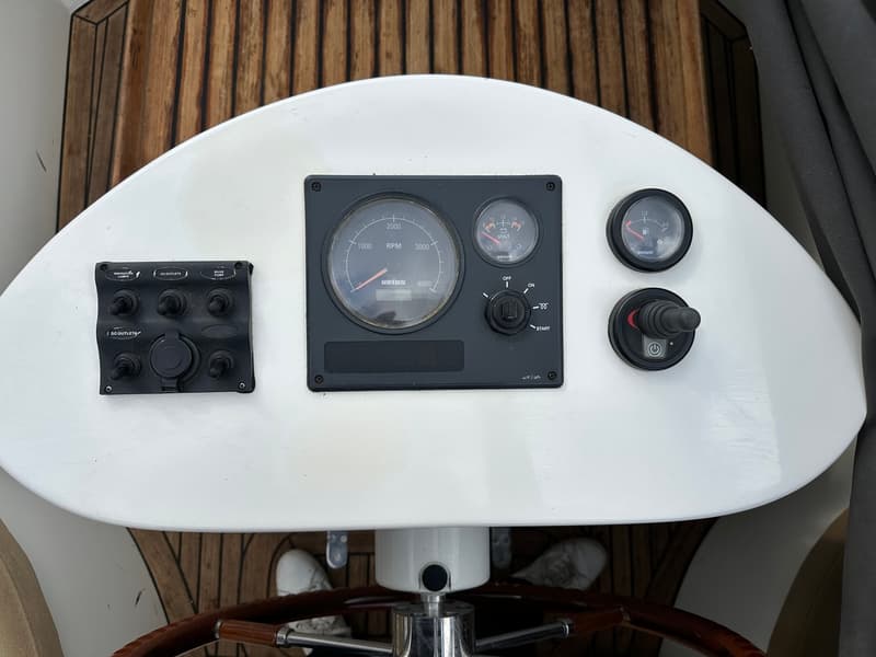 Een wit dashboard met diverse meters en schakelaars, met een houten vloer en een stuurwiel eronder.