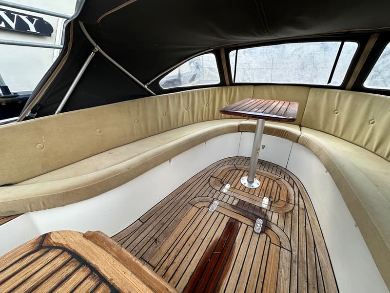 Een bootinterieur met een houten vloer, beige kussens en een houten tafel onder een zwarte kap.