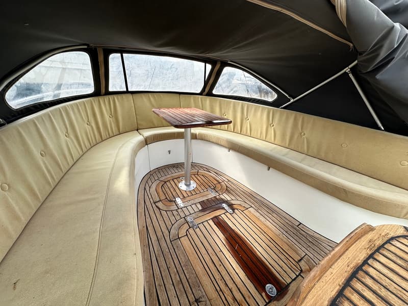 De foto toont een bootinterieur met een U-vormige zitbank met beige kussens, een houten tafel en een houten vloer.