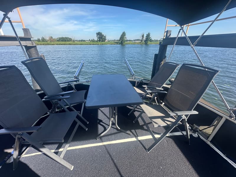 Een dek met een tafel en vier stoelen, met uitzicht op water en een groene oever.