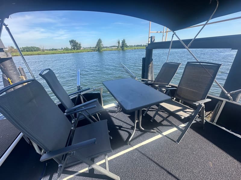 Een dek met vier donkere stoelen en een donkere tafel, met uitzicht op water en een groene oever.