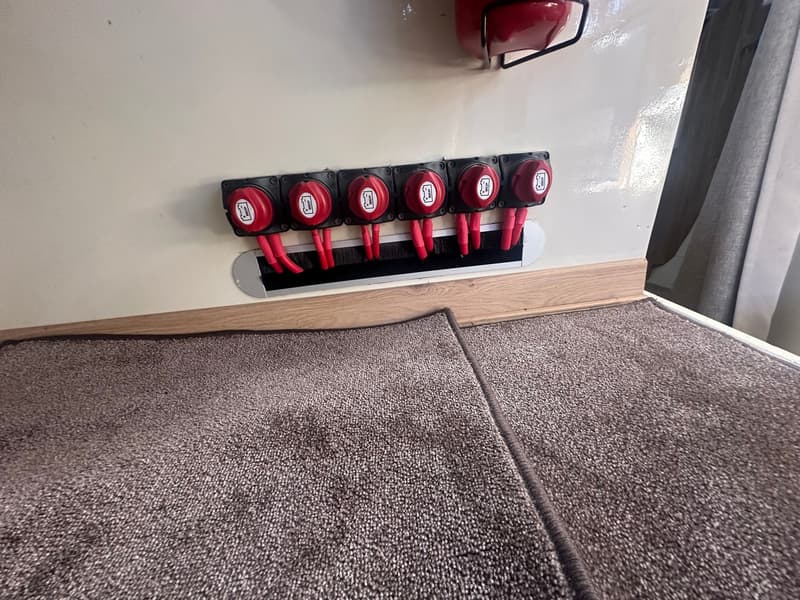 Zes rode hoofdschakelaars met rode kabels zijn gemonteerd op een witte wand boven een grijze vloerbedekking.